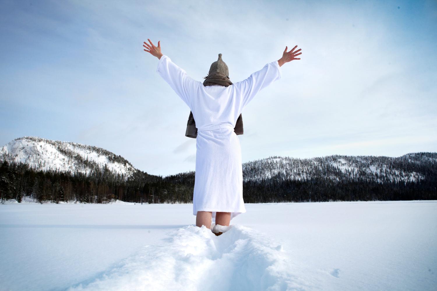 Henkilö seisoo lumihangessa kylpytakki ja saunahattu päällään, kädet ylhäällä ja selkä kameraan päin.
