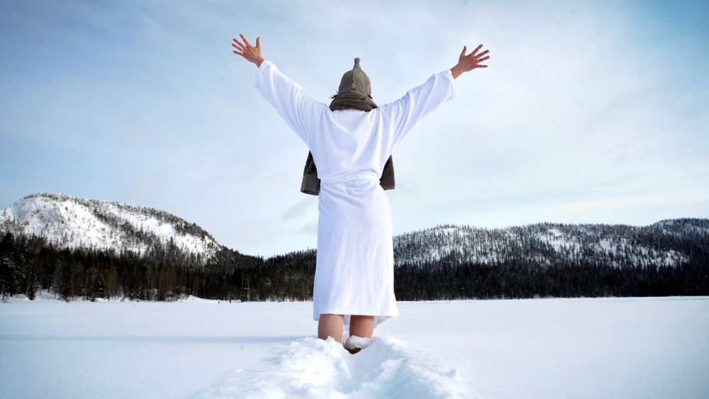 A person throws their hands in the air and stands in the snow wearing a bathrobe and a sauna hat.